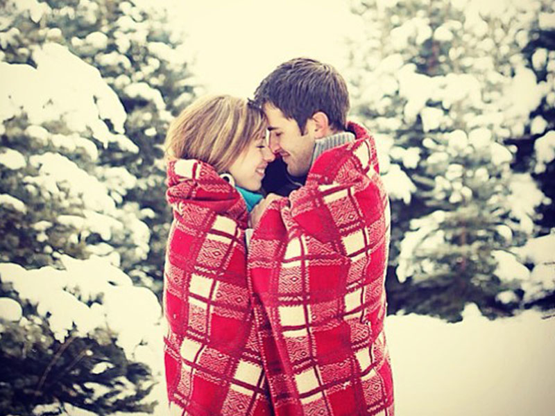 A couple in the snow wrapped up in a blanket together snuggling.