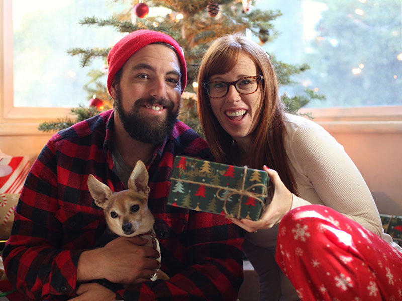 A couple in a healthy relationship in front of a Christmas tree laughing with a dog.