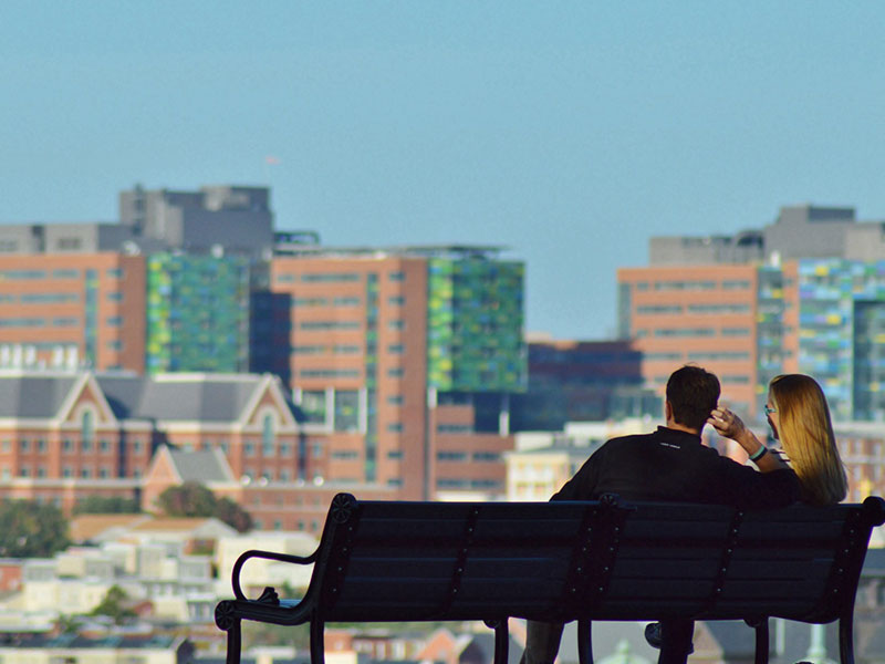 A woman and a man in their 30s on a park bench looking at a city on a date.