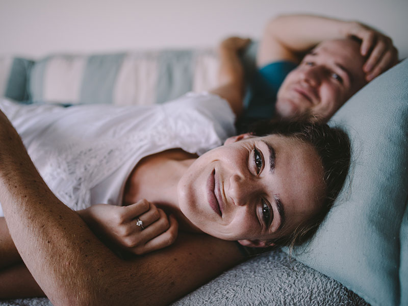 A woman who read the signs that he still loves you and is in bed smiling with her boyfriends arm around her.
