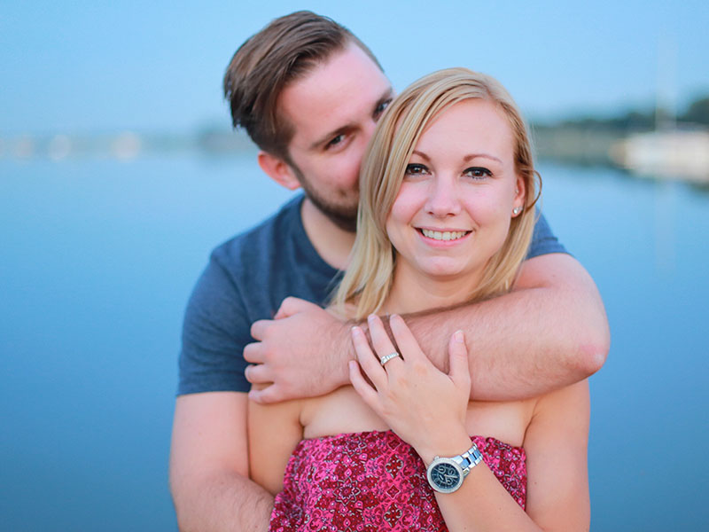 A woman who's dating an introvert smiling while her boyfriend stands behind her hiding and kissing her hair.