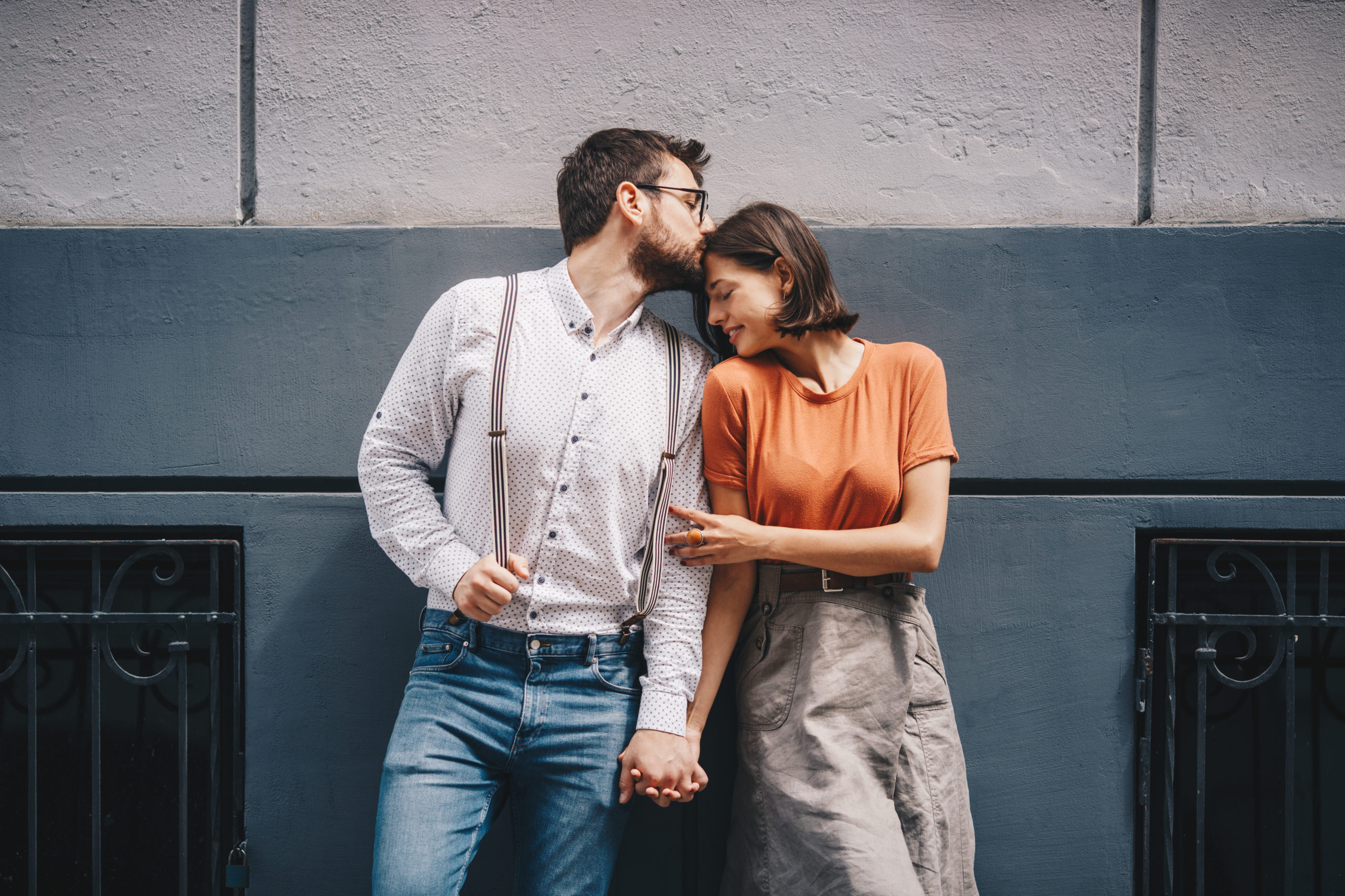 Happy couple standing outdoors with backs against a wall while man kisses woman on the head as they're in a lust vs. love relationship.