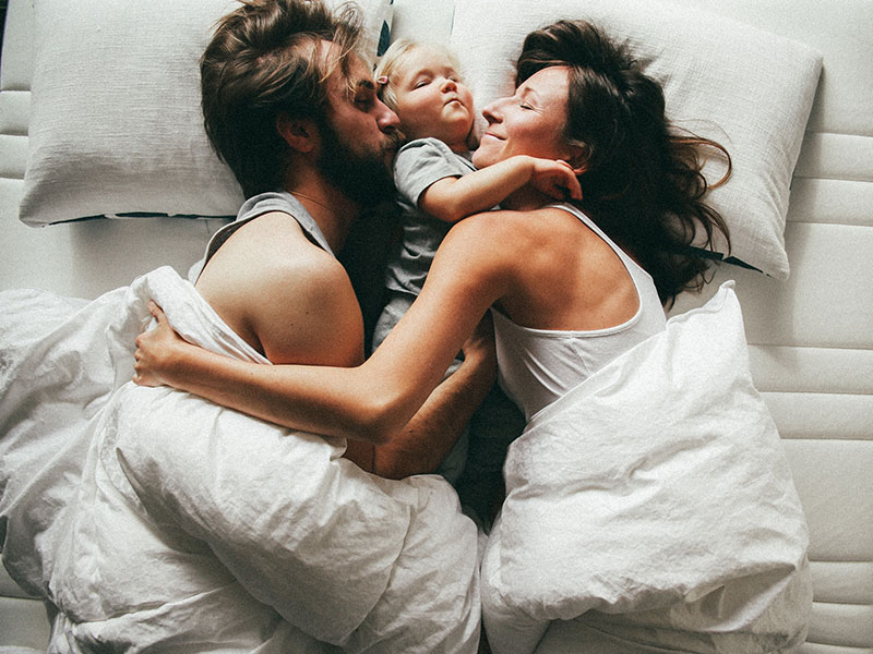 A couple who practices co-leading in their marriage in bed together laughing with their child.