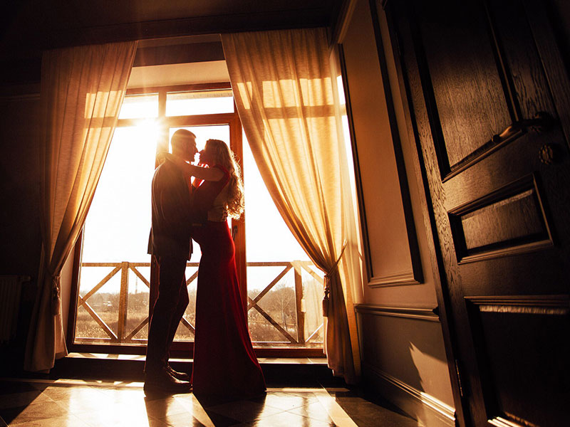 A hopeless romantic couple kissing in front of a window at sunset.