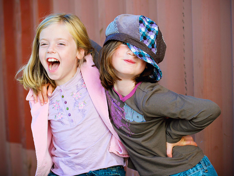 Two little girls who are childhood friends laughing and joking around together in front of a fence.