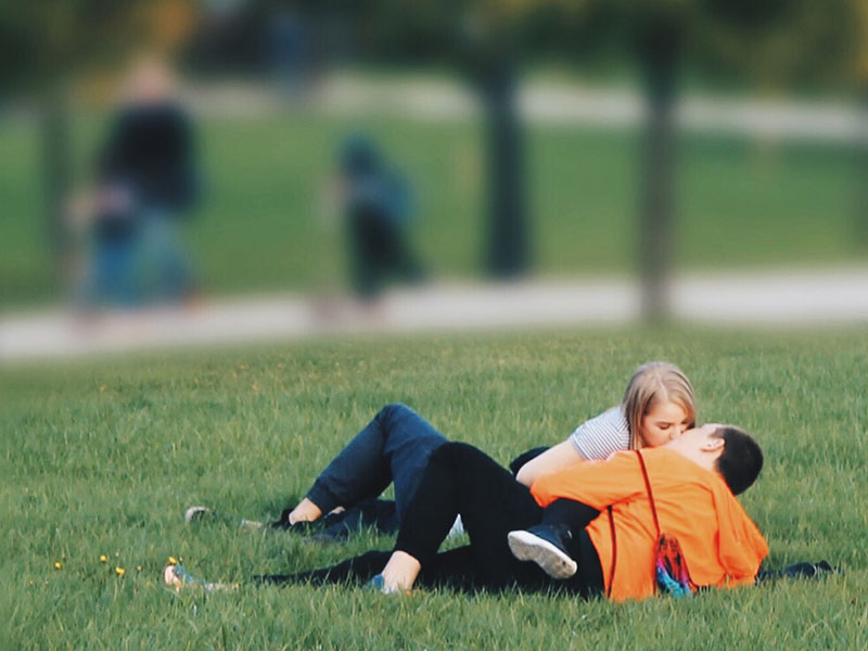 A couple dating in college, kissing in the grass in the middle of campus.