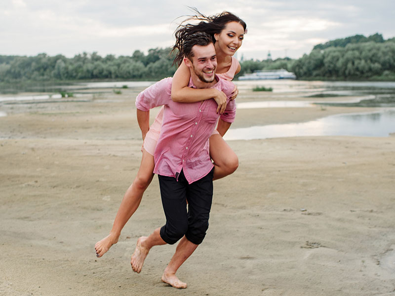 A girl wondering "does he like me" smiling and laughing as a guy friend carries her on his back on the beach.