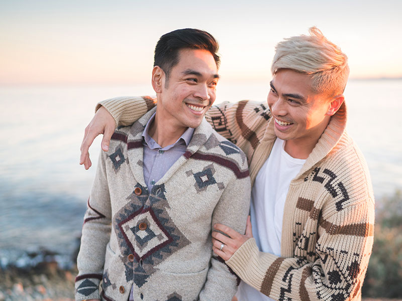 A gay couple on the beach laughing together as they take a photo for social media sites.
