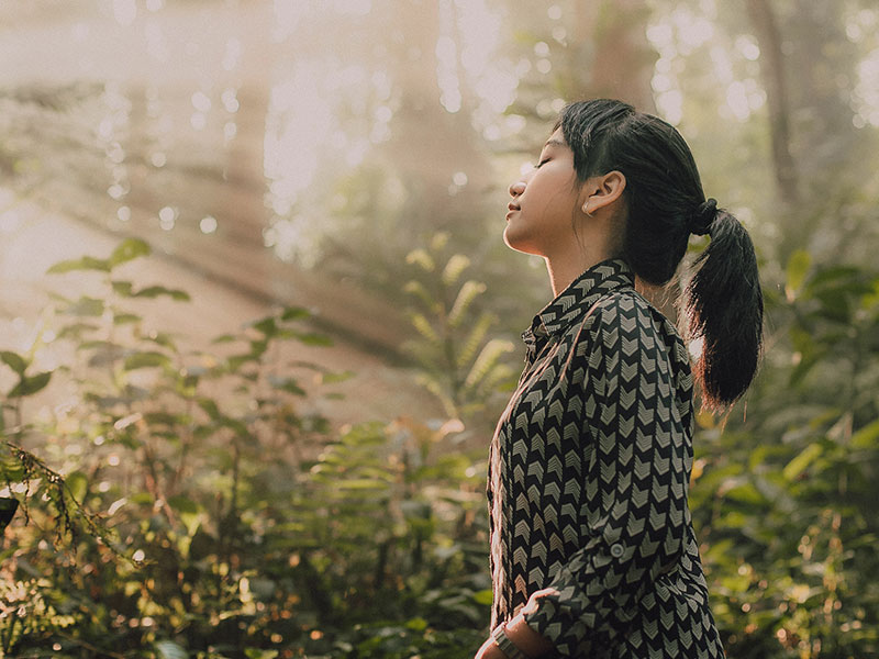 A woman who learned that sometimes you have to let go, standing in the forest with her eyes closed breathing in the fresh air.