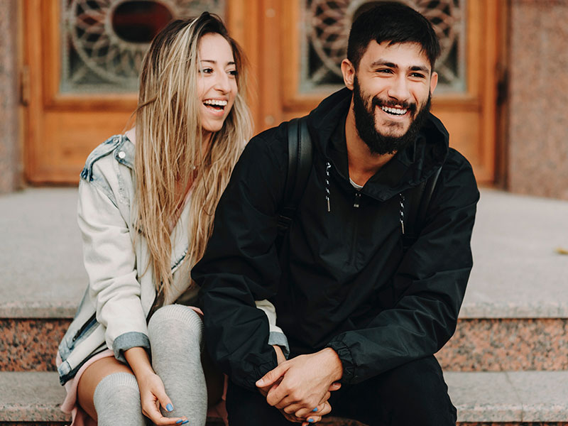 A guy who learned how to impress a girl laughing with his girlfriend who's smiling at him and holding his hand.