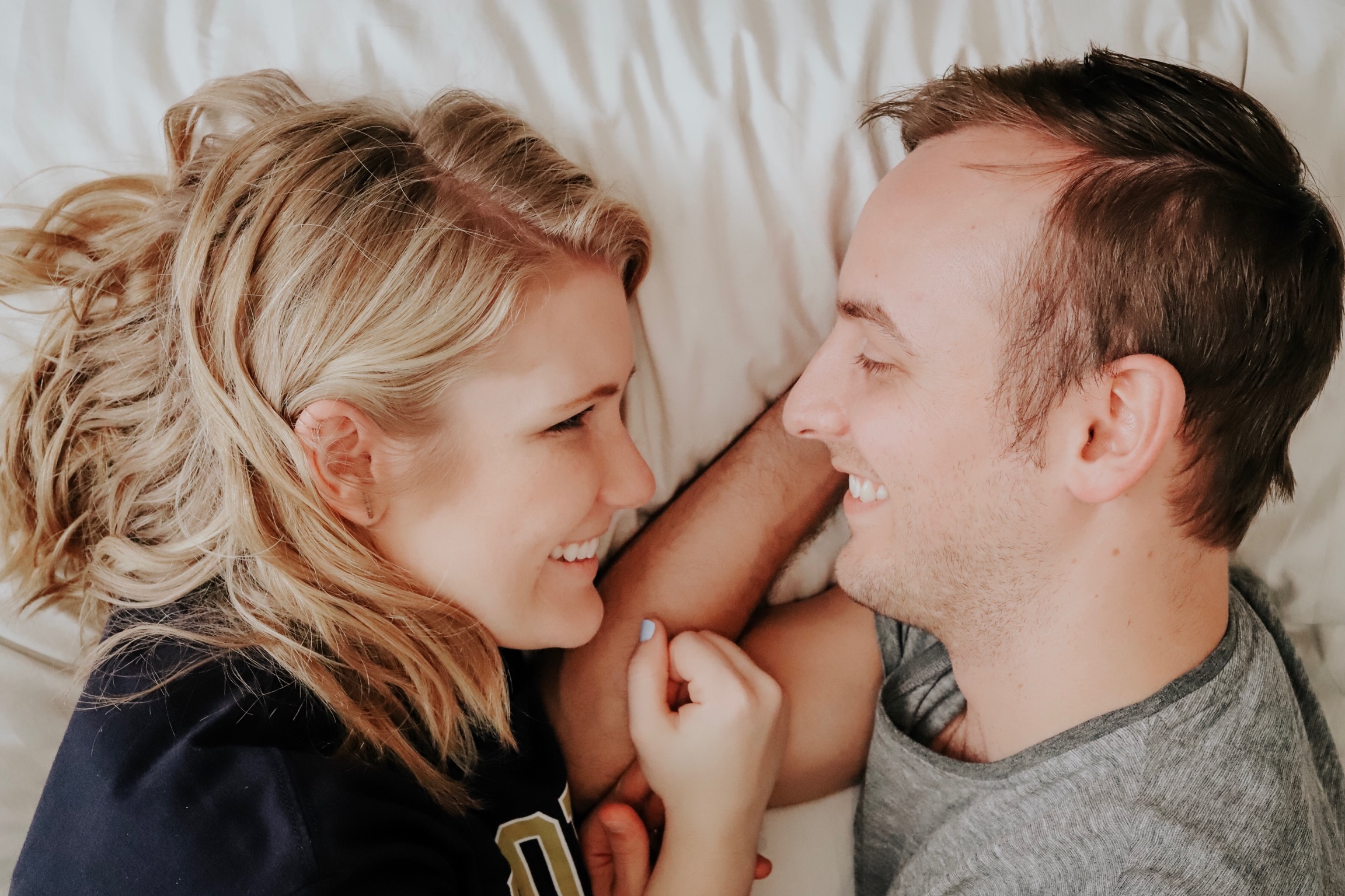 Happy couple lying in bed looking at each other and smiling while talking about winter sex ideas.