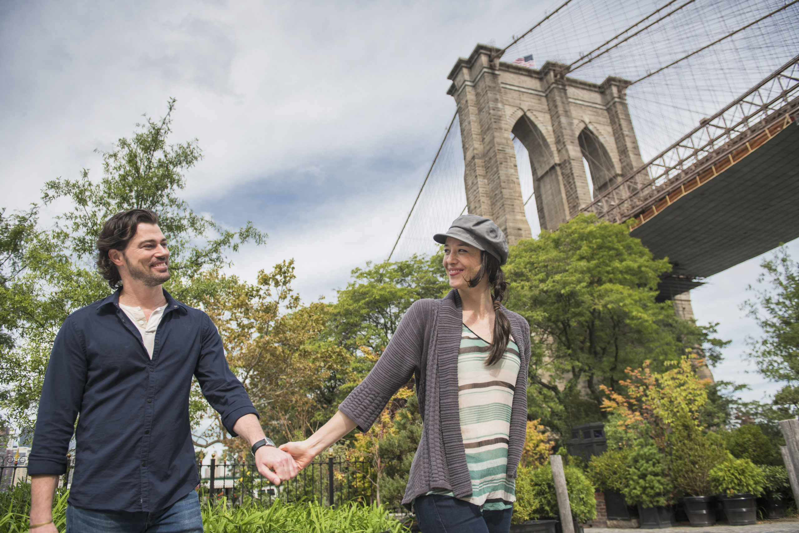 Happy couple holding hands and walking with Brooklyn Bridge in background while trying out some of the best date ideas in NYC.